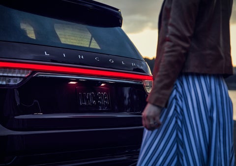 A person is shown near the rear of a 2024 Lincoln Aviator® SUV as the Lincoln Embrace illuminates the rear lights | White's Canyon Motors - Lincoln in Spearfish SD